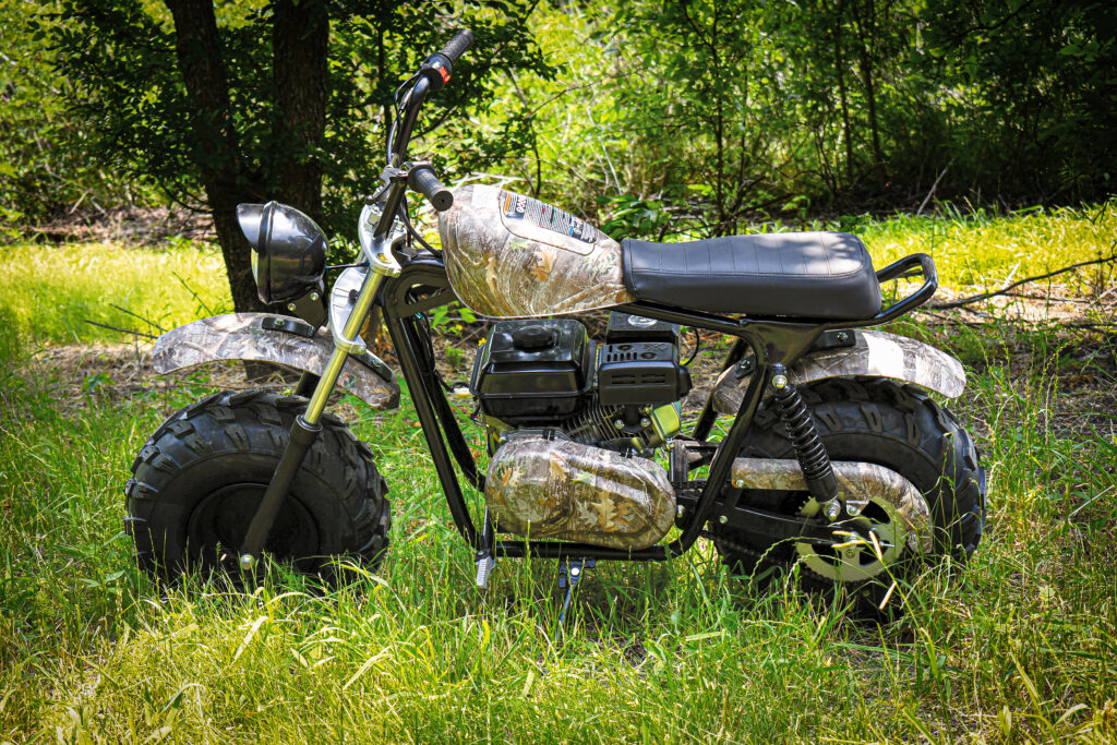 Camo-patterned mini bike with oversized tires parked in a grassy woodland area.